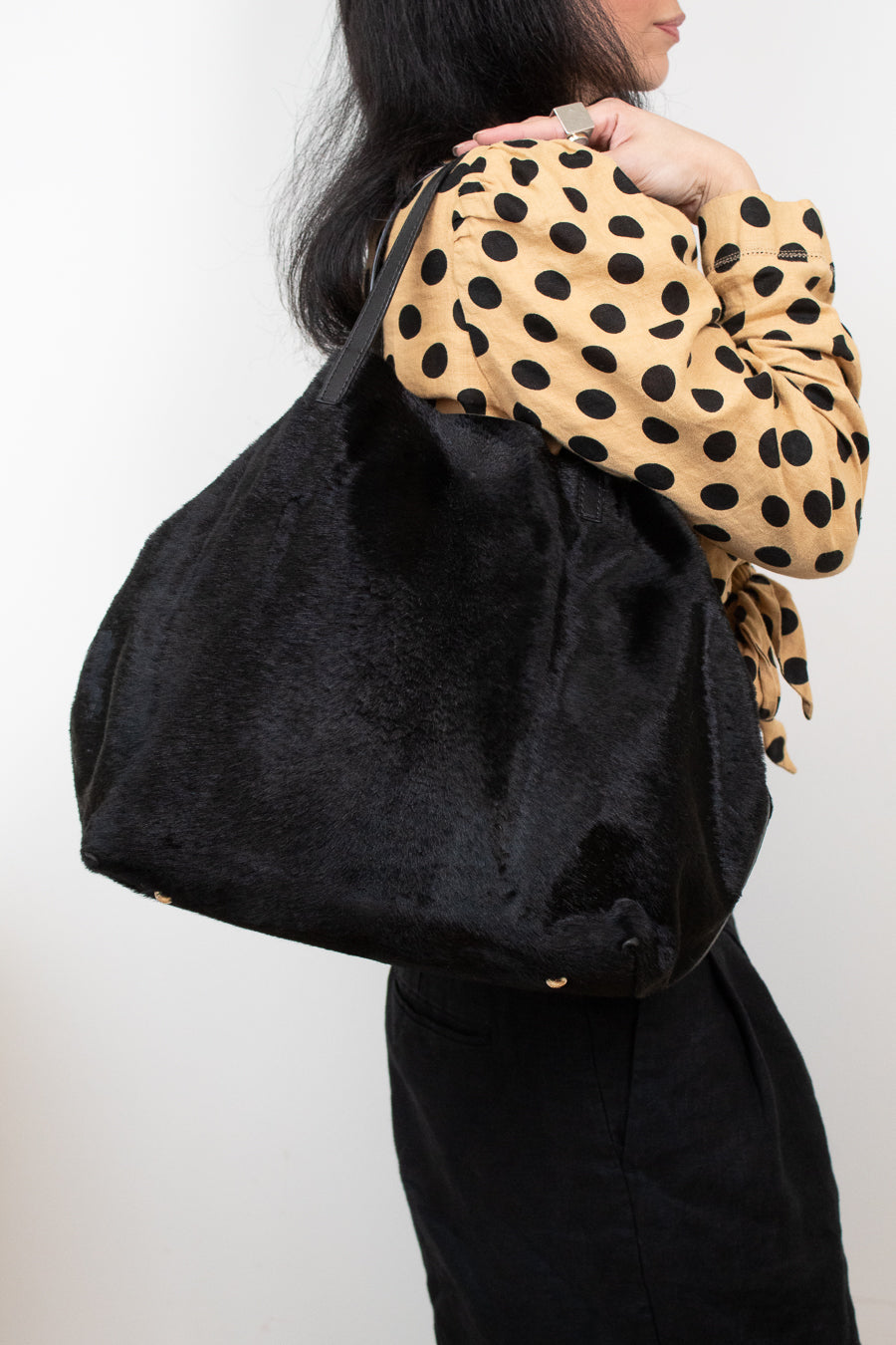 A person wearing a tan and black polka dot blouse carries a large black fuzzy shoulder bag against a plain light background.