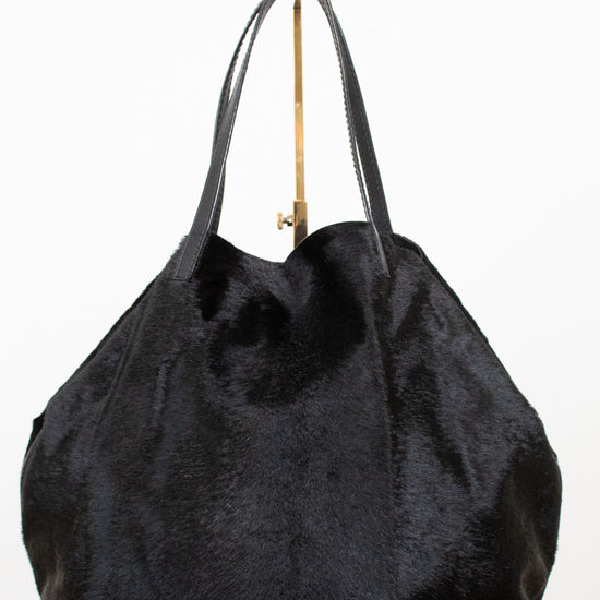 A black faux fur tote bag with black leather shoulder straps and a gold-tone metal hanger, shown against a plain white background.