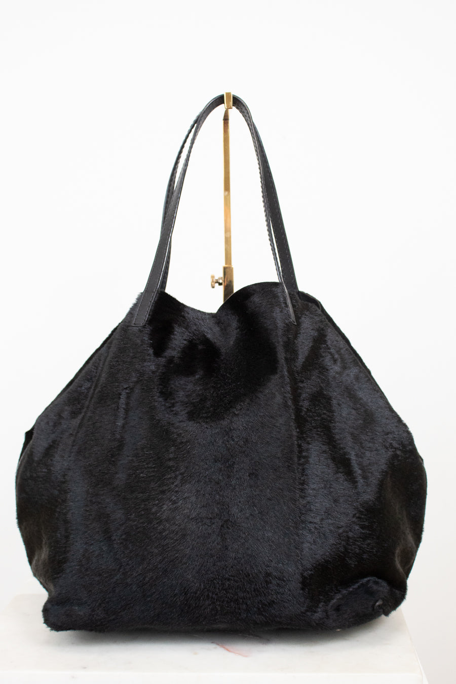 A black faux fur tote bag with black leather shoulder straps and a gold-tone metal hanger, shown against a plain white background.