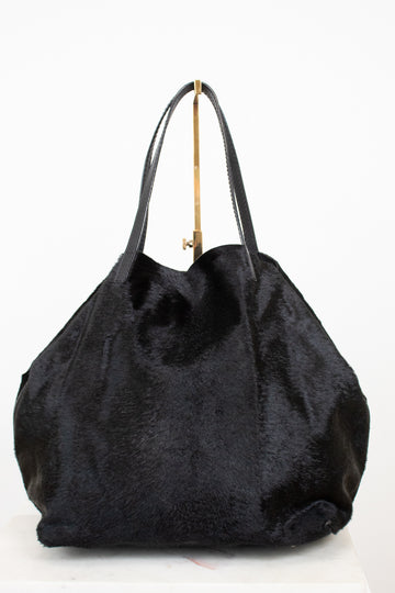 A black faux fur tote bag with black leather shoulder straps and a gold-tone metal hanger, shown against a plain white background.