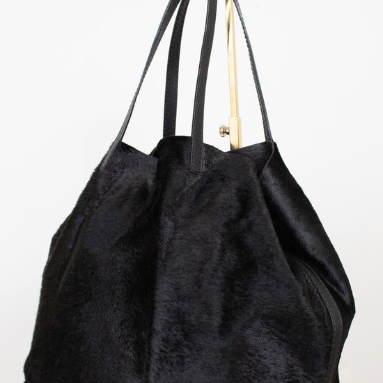 A black textured faux fur or shearling-style tote bag with long shoulder straps  photographed on a plain white studio background.