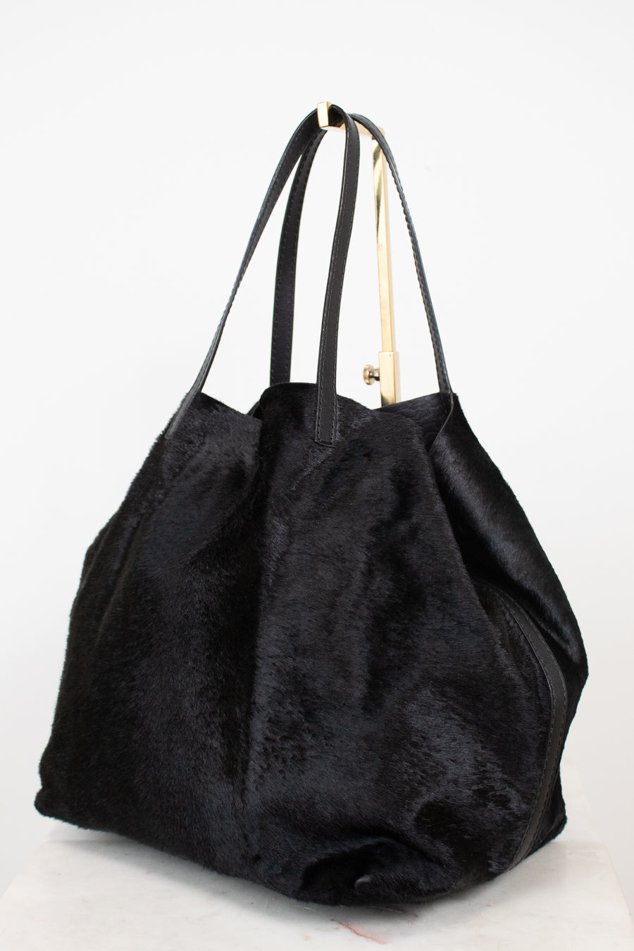 A black textured faux fur or shearling-style tote bag with long shoulder straps  photographed on a plain white studio background.
