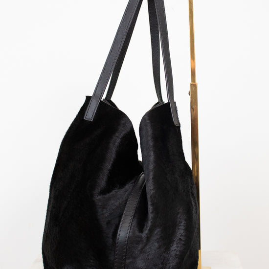A black fuzzy slouchy tote bag with dark gray shoulder straps, displayed on a gold metal stand against a white background.