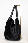 A black fuzzy slouchy tote bag with dark gray shoulder straps, displayed on a gold metal stand against a white background.