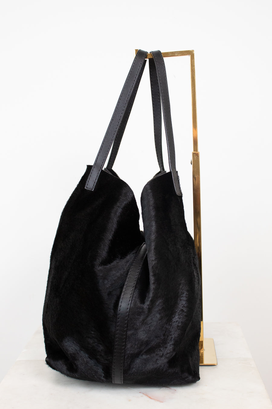 A black fuzzy slouchy tote bag with dark gray shoulder straps, displayed on a gold metal stand against a white background.