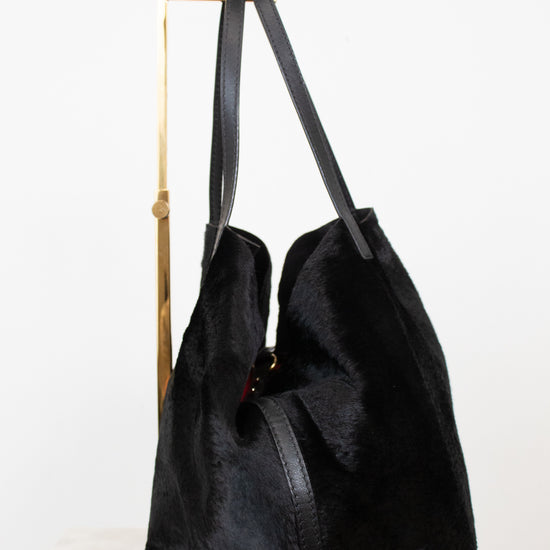 A black faux fur  tote bag with black leather shoulder straps, displayed on a gold metal stand against a white background.