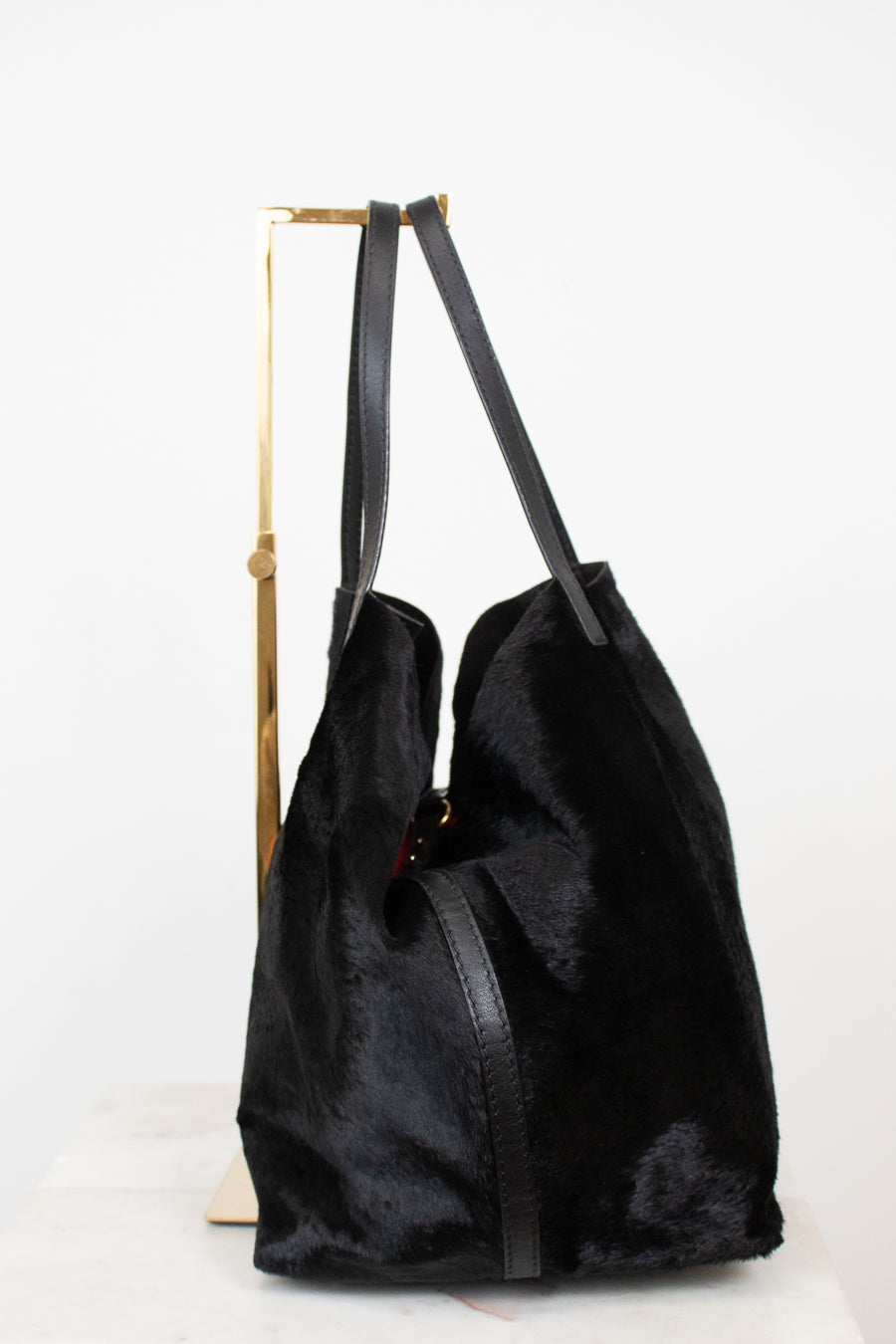 A black faux fur  tote bag with black leather shoulder straps, displayed on a gold metal stand against a white background.