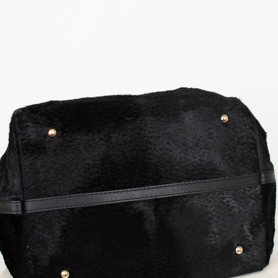 A black faux fur bag with a black leather strap and small gold-tone metal studs, resting on a white marble surface against a white background.