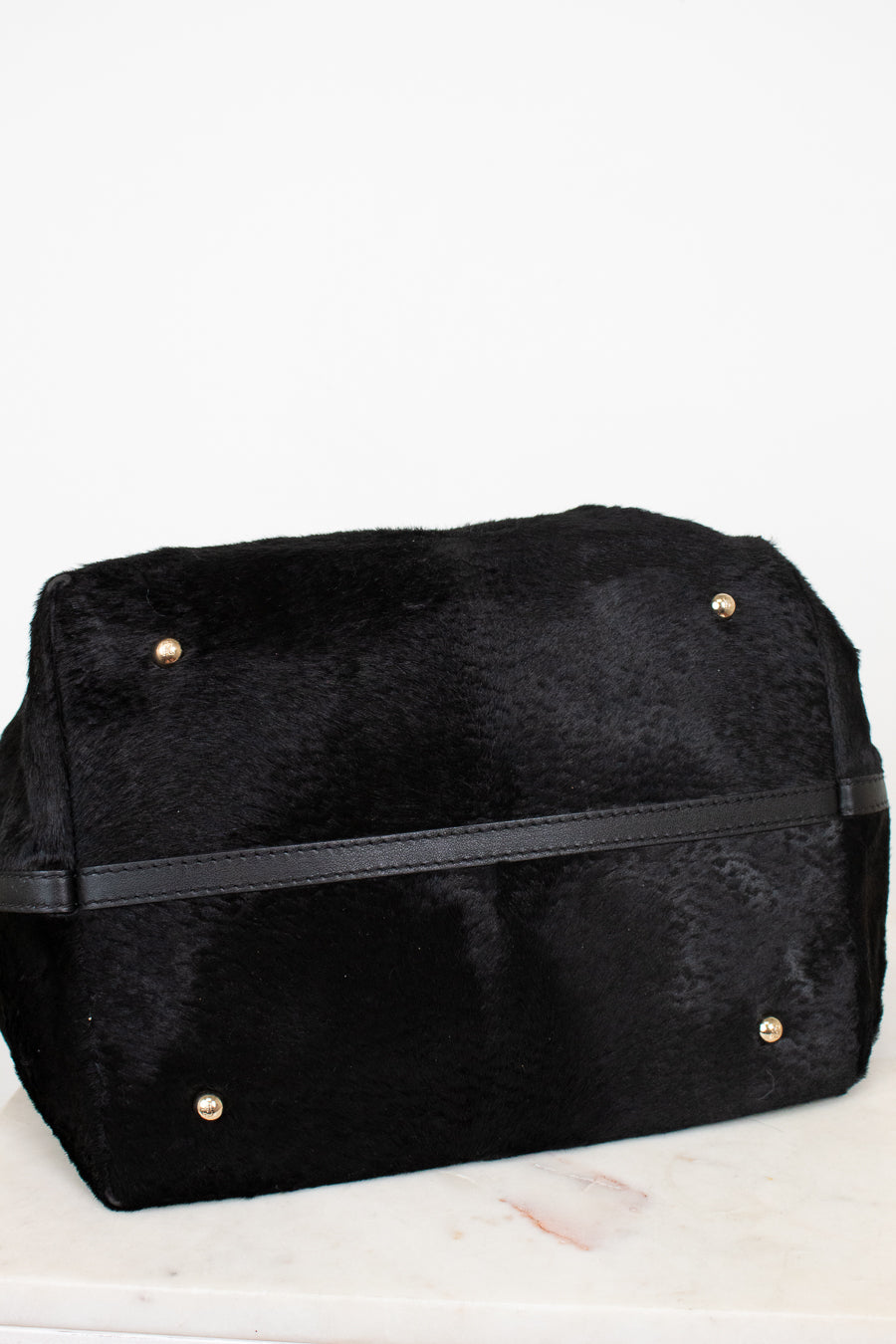 A black faux fur bag with a black leather strap and small gold-tone metal studs, resting on a white marble surface against a white background.