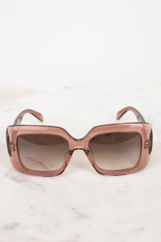 Oversized square sunglasses with a translucent dusty rose pink frame and gradient brown lenses, resting on a light marble surface against a white background.