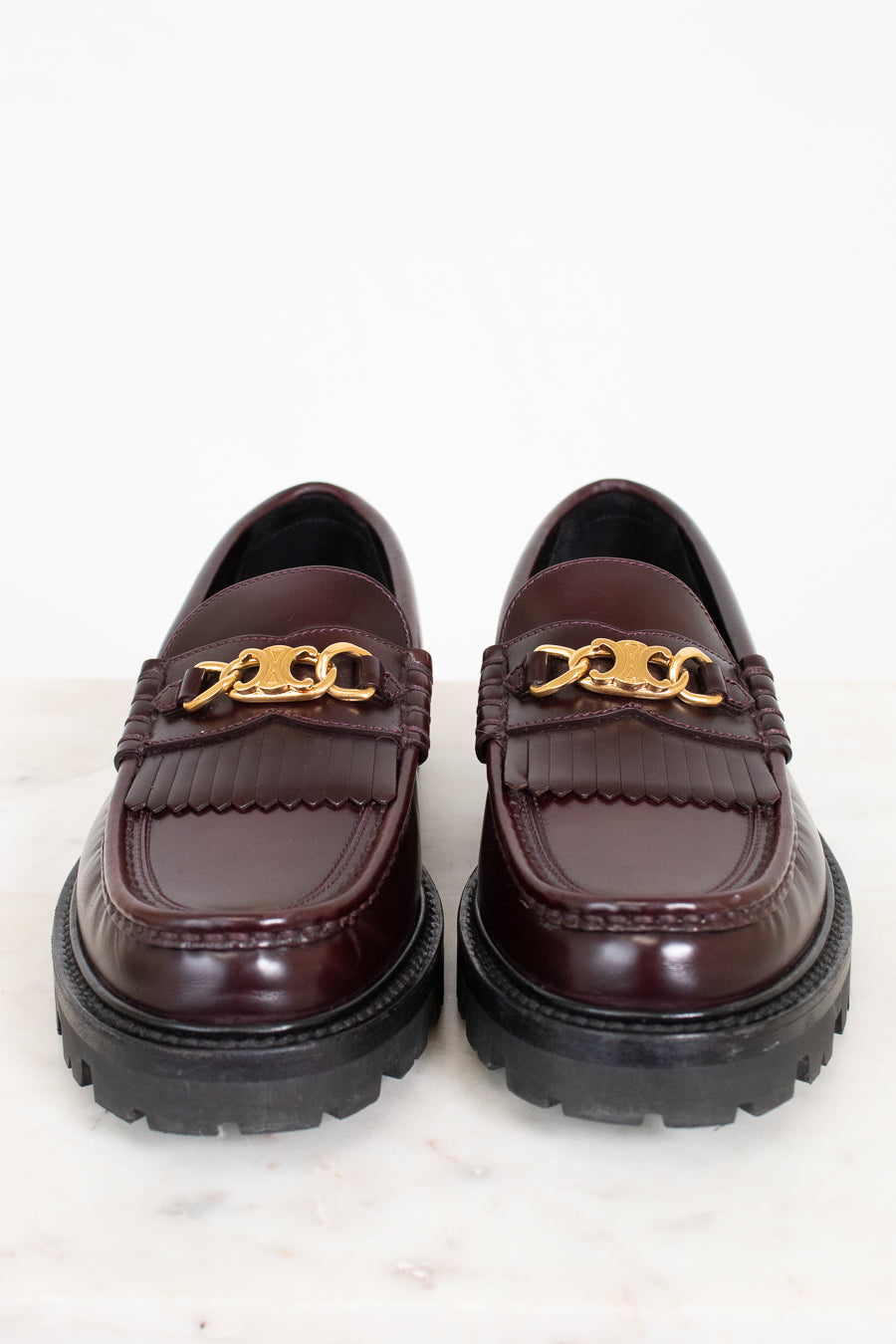 Pair of brown loafers with gold chain detail on a white background