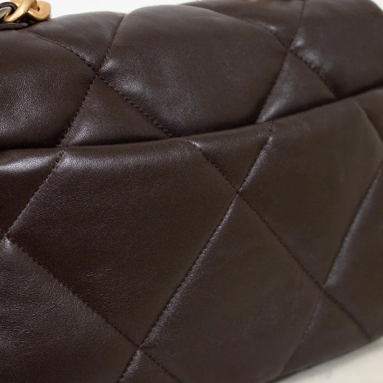 Close-up of a dark brown quilted handbag with gold chain handle on a light marble surface.