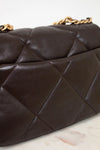 Close-up of a dark brown quilted handbag with gold chain handle on a light marble surface.
