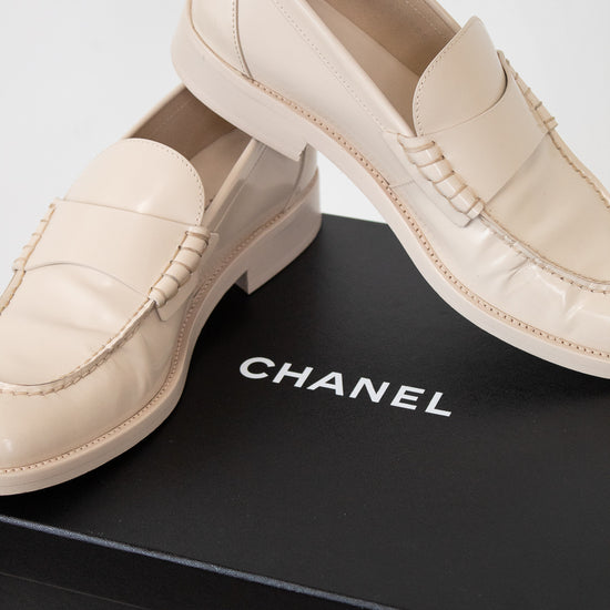 A pair of Chanel loafers in light beige leather, displayed on top of a black Chanel shoebox against a clean white background.