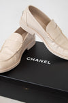 A pair of Chanel loafers in light beige leather, displayed on top of a black Chanel shoebox against a clean white background.