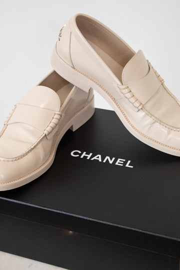 A pair of Chanel loafers in light beige leather, displayed on top of a black Chanel shoebox against a clean white background.