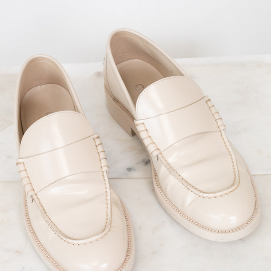 A pair of cream-colored leather loafers with decorative stitching, displayed on a white marble surface against a white background.