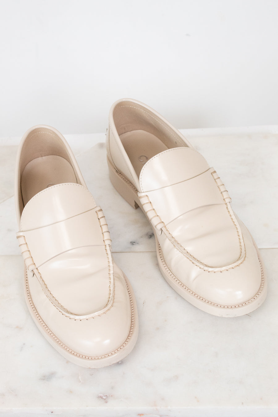 A pair of cream-colored leather loafers with decorative stitching, displayed on a white marble surface against a white background.