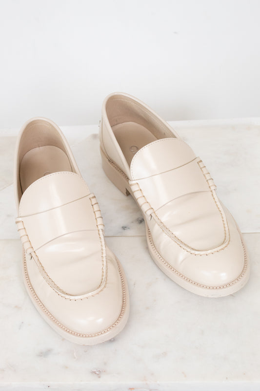A pair of cream-colored leather loafers with decorative stitching, displayed on a white marble surface against a white background.