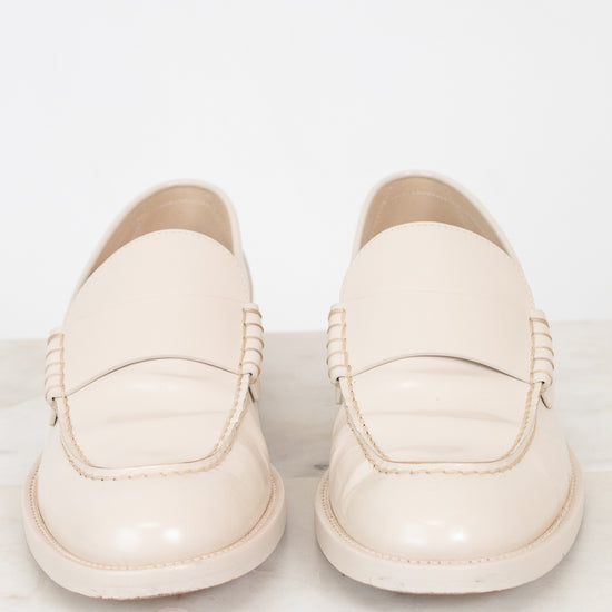 A pair of cream-colored leather loafers with decorative braided detailing on the sides, displayed on a light marble surface against a white background.