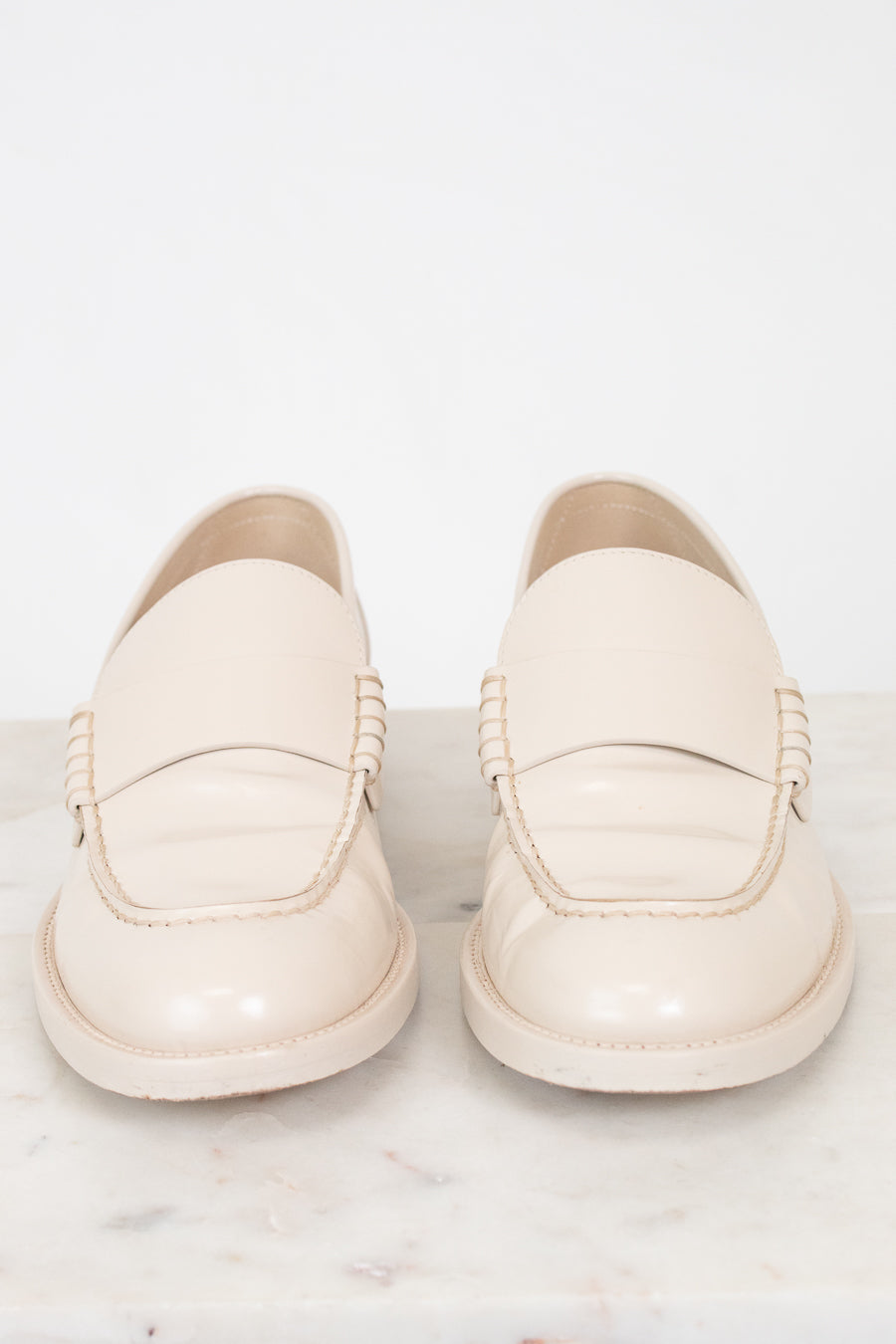 A pair of cream-colored leather loafers with decorative braided detailing on the sides, displayed on a light marble surface against a white background.