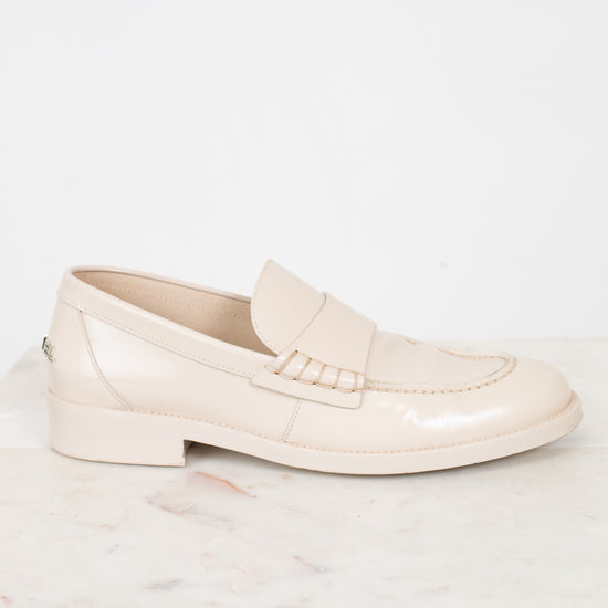 A single light beige patent leather loafer shoe resting on a white marble slab against a white background.