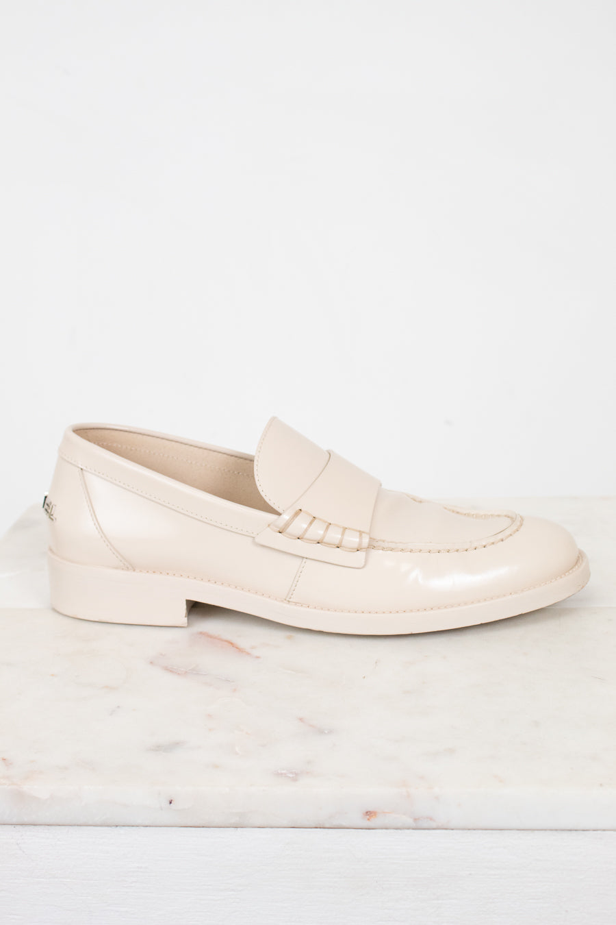 A single light beige patent leather loafer shoe resting on a white marble slab against a white background.