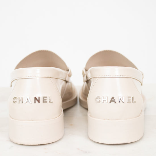 A pair of Chanel loafers shoes in a light cream or beige patent leather, shown from the back on a white marble surface against a white background.