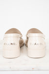 A pair of Chanel loafers shoes in a light cream or beige patent leather, shown from the back on a white marble surface against a white background.