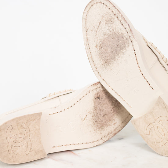 A pair of beige leather Chanel loafers shown from the underside, resting on a white marble surface against a white background. The soles feature the embossed Chanel logo and show some signs of wear.