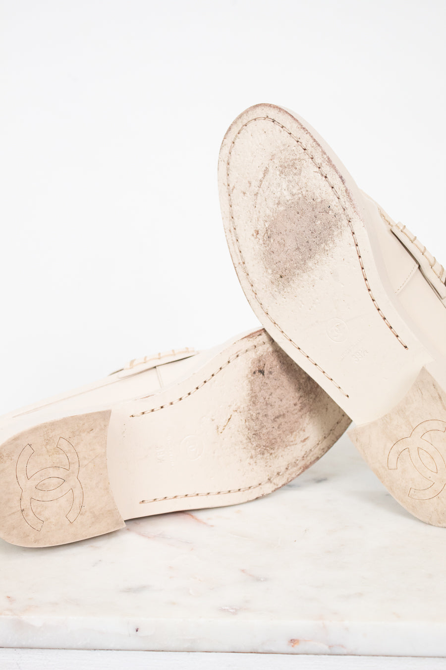 A pair of beige leather Chanel loafers shown from the underside, resting on a white marble surface against a white background. The soles feature the embossed Chanel logo and show some signs of wear.