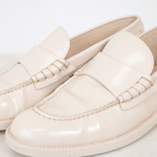 A pair of glossy cream-colored loafers with decorative stitching on the strap, displayed on a light marble surface against a white background.