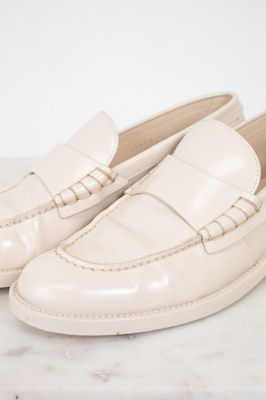 A pair of glossy cream-colored loafers with decorative stitching on the strap, displayed on a light marble surface against a white background.