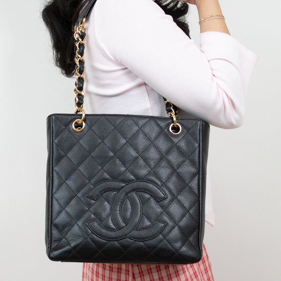 A black quilted Chanel tote bag with gold chain straps and a gold interlocking CC logo, shown being worn by a model in a white top and red-and-white gingham skirt against a plain light background.