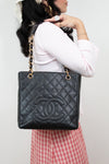 A black quilted Chanel tote bag with gold chain straps and a gold interlocking CC logo, shown being worn by a model in a white top and red-and-white gingham skirt against a plain light background.