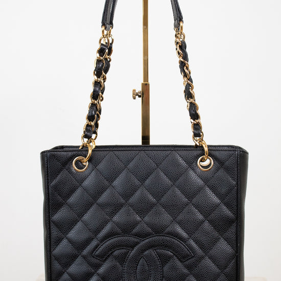 A black quilted leather Chanel tote bag with a large embossed CC logo on the front, featuring gold-tone chain and leather shoulder straps, displayed on a stand against a white background.