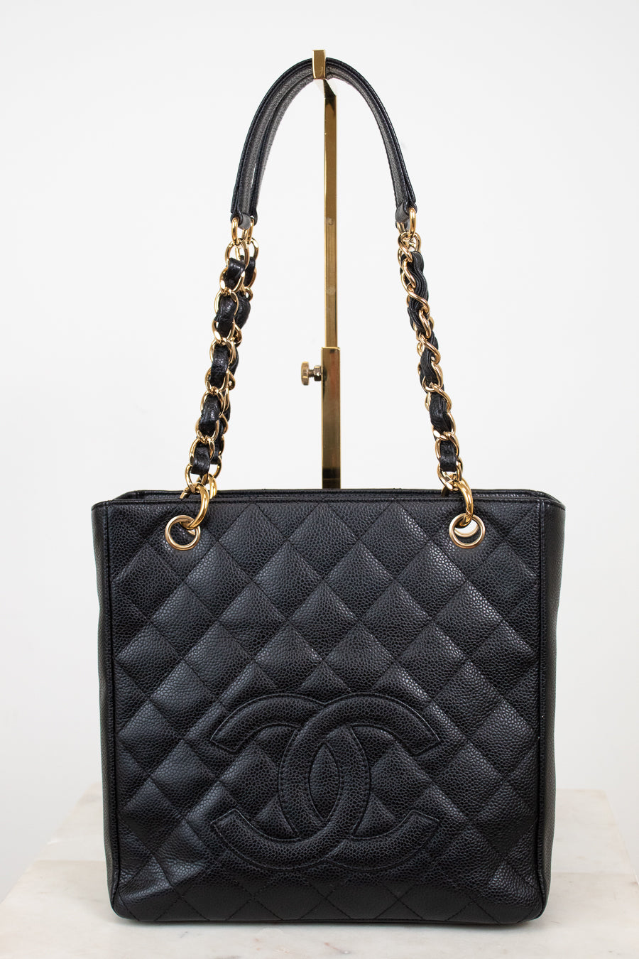 A black quilted leather Chanel tote bag with a large embossed CC logo on the front, featuring gold-tone chain and leather shoulder straps, displayed on a stand against a white background.