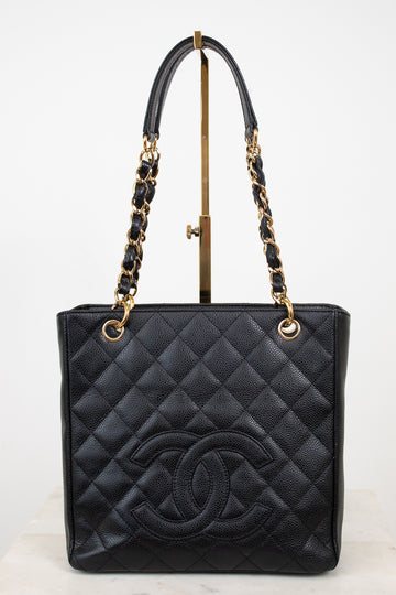 A black quilted leather Chanel tote bag with a large embossed CC logo on the front, featuring gold-tone chain and leather shoulder straps, displayed on a stand against a white background.