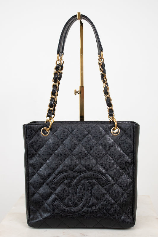 A black quilted leather Chanel tote bag with a large embossed CC logo on the front, featuring gold-tone chain and leather shoulder straps, displayed on a stand against a white background.