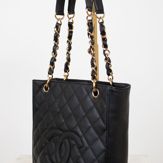 A black quilted leather tote bag with gold-tone hardware, featuring a large embossed interlocking CC logo on the front and dual top handles, shown on a light neutral background.