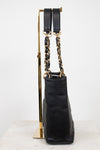 A black quilted leather handbag with gold-tone chain and strap hardware, displayed upright on a gold stand against a white studio background.