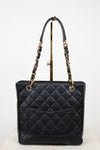 A black quilted leather tote bag with gold-tone chain handles and gold hardware, displayed on a stand against a white studio background.