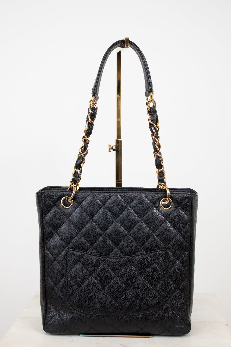 A black quilted leather tote bag with gold-tone chain handles and gold hardware, displayed on a stand against a white studio background.