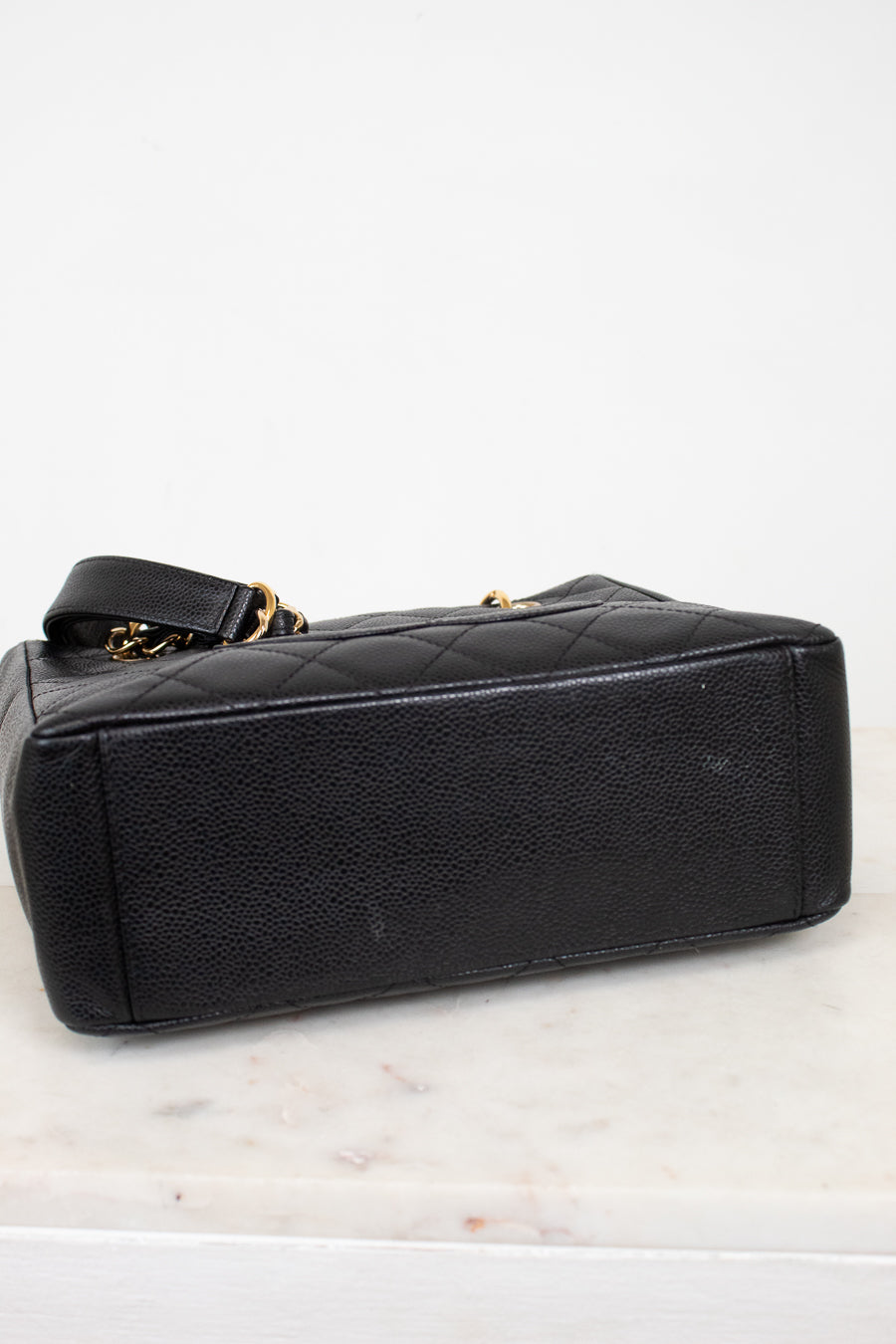 A black quilted leather handbag with gold-tone chain accents, shown from a side angle on a white marble surface against a white background.