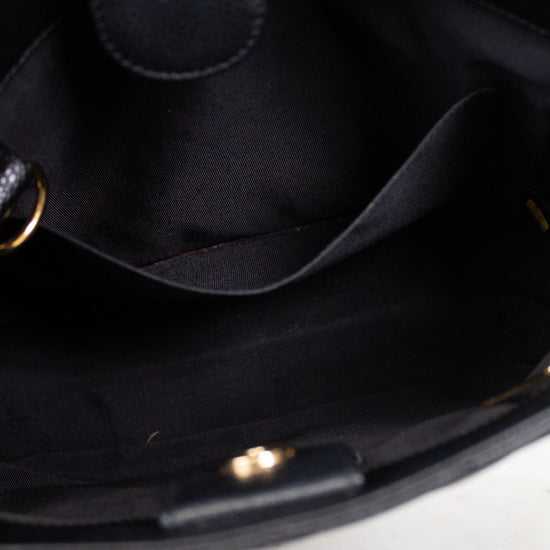 Close-up of the interior of a black handbag, showing a black fabric lining, interior pocket, and small gold-tone hardware. A hand with lavender nail polish is partially visible at the top edge, resting on a light marble surface.