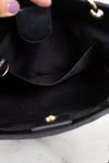 Close-up of the interior of a black handbag, showing a black fabric lining, interior pocket, and small gold-tone hardware. A hand with lavender nail polish is partially visible at the top edge, resting on a light marble surface.