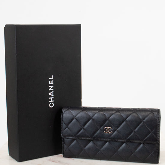 A black Chanel quilted leather wallet with a gold CC turn-lock, displayed next to its black Chanel box on a light marble surface against a white background.