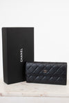 A black Chanel quilted leather wallet with a gold CC turn-lock, displayed next to its black Chanel box on a light marble surface against a white background.