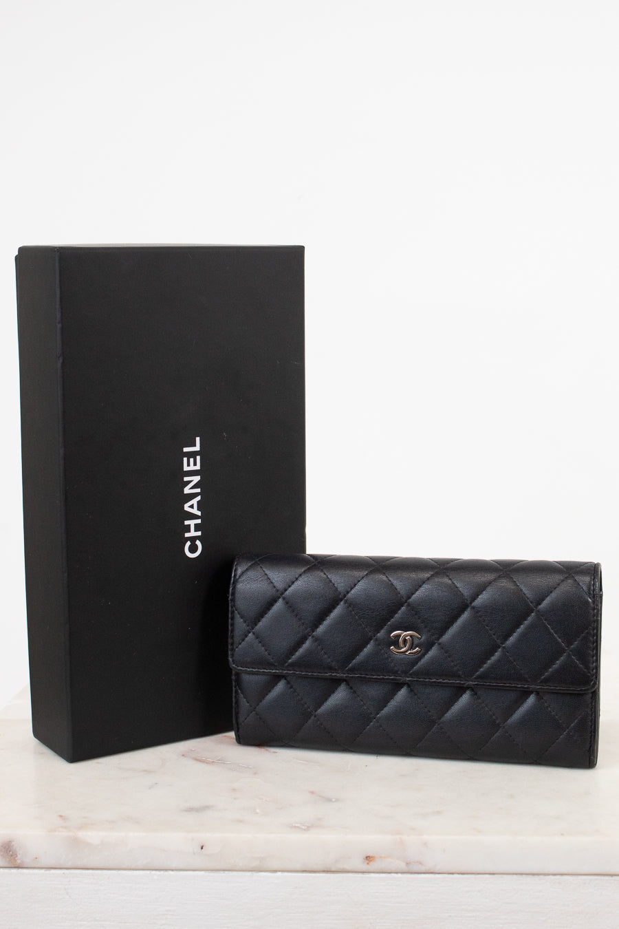 A black Chanel quilted leather wallet with a gold CC turn-lock, displayed next to its black Chanel box on a light marble surface against a white background.