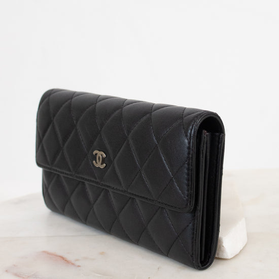 A black quilted leather wallet with a silver-tone interlocking CC logo, displayed on a white marble surface against a white background.